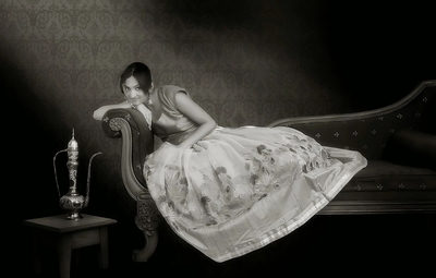 A graceful black-and-white portrait featuring a woman in a traditional embroidered dress reclining on an ornate vintage sofa. Soft lighting, classic décor, and delicate textures create an elegant, timeless atmosphere with a touch of old-world charm.